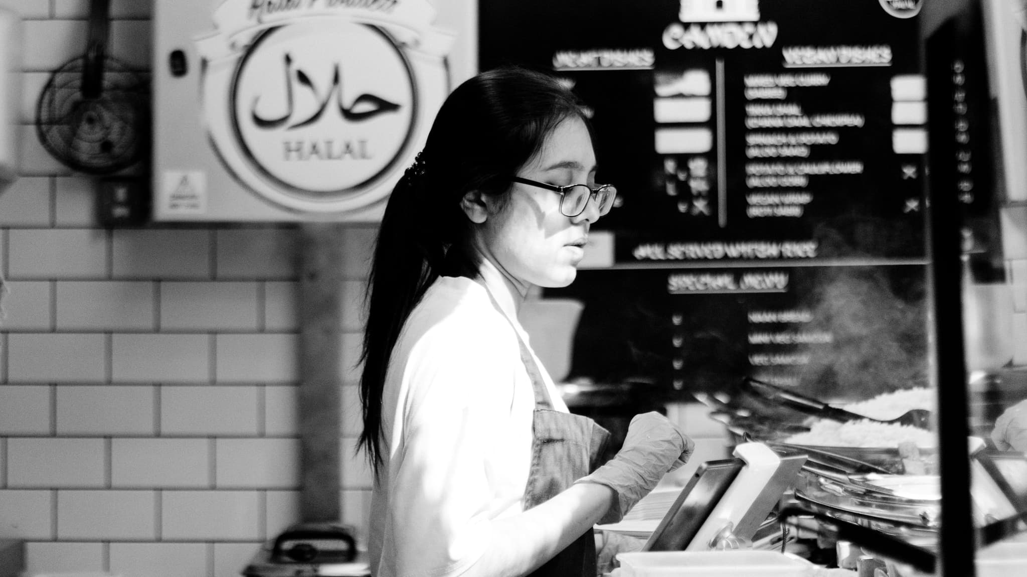 A black and white photo captures a person working behind the counter at a food establishment with a menu board in the background, indicating Halal options available