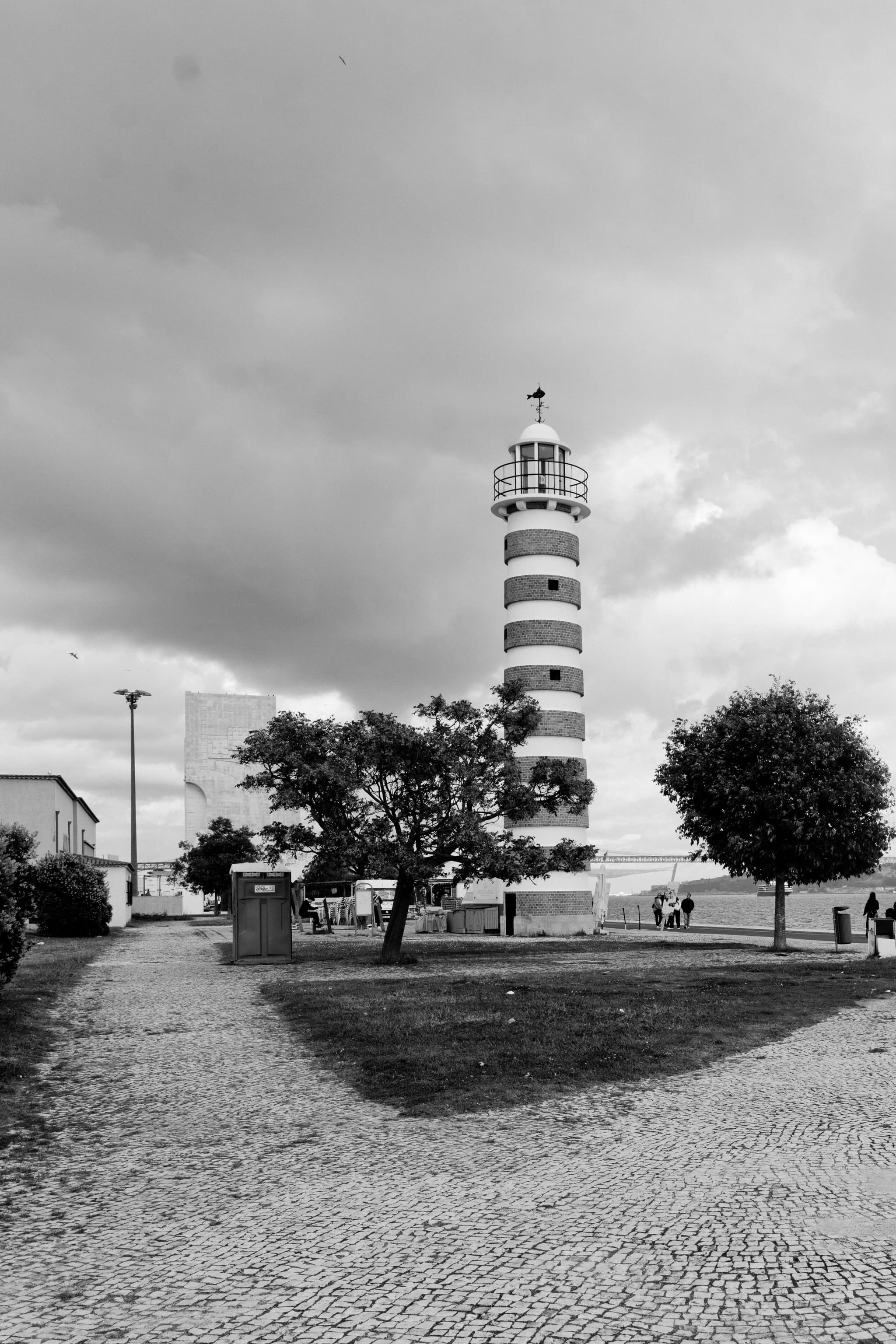 A black and white photograph of a striped lighthouse surrounded by trees and a cobblestone path, with a cloudy sky overhead