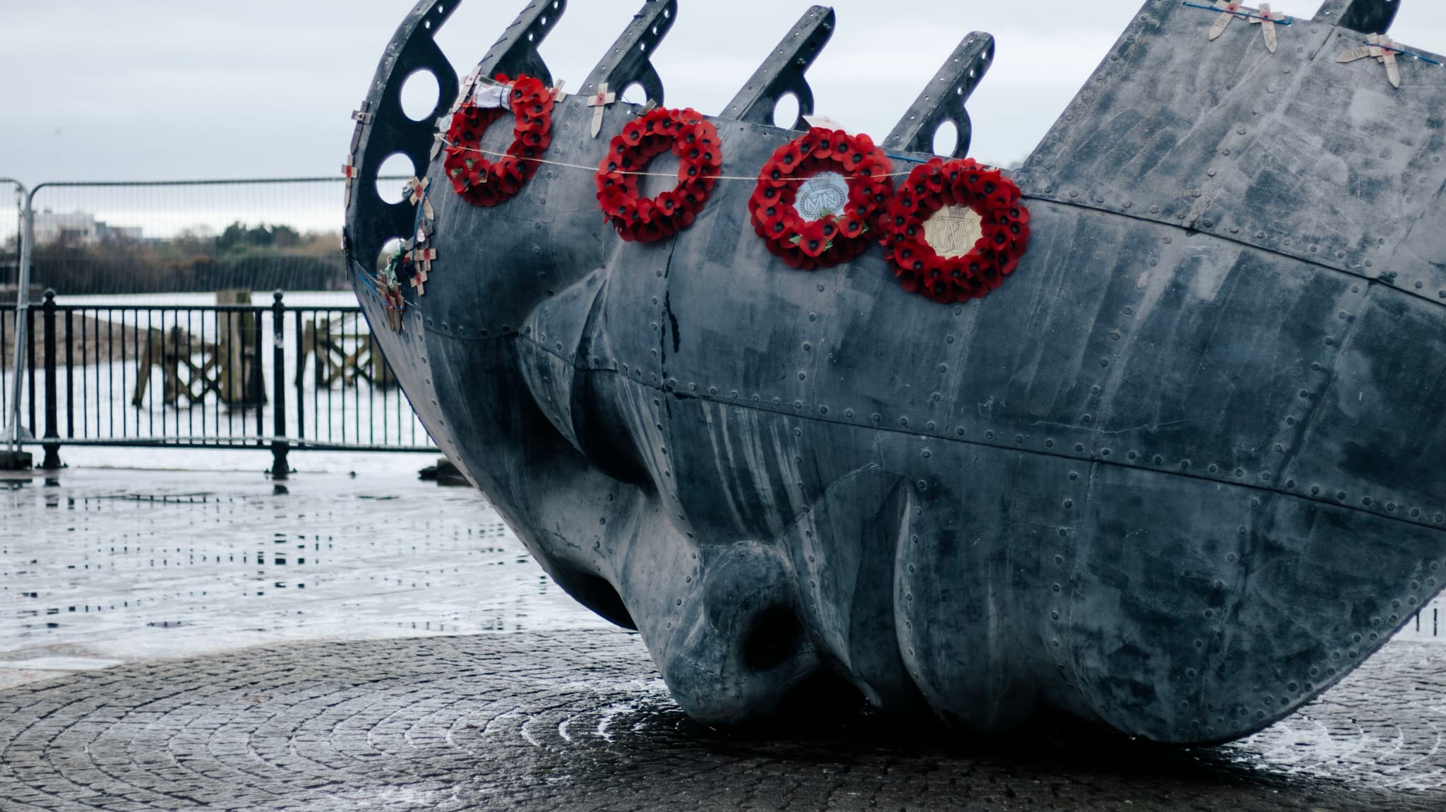 A gray, weathered metal sculpture resembling a ship's hull adorned with several red wreaths, positioned in an outdoor setting with a wet ground, suggesting a commemorative installation or monument