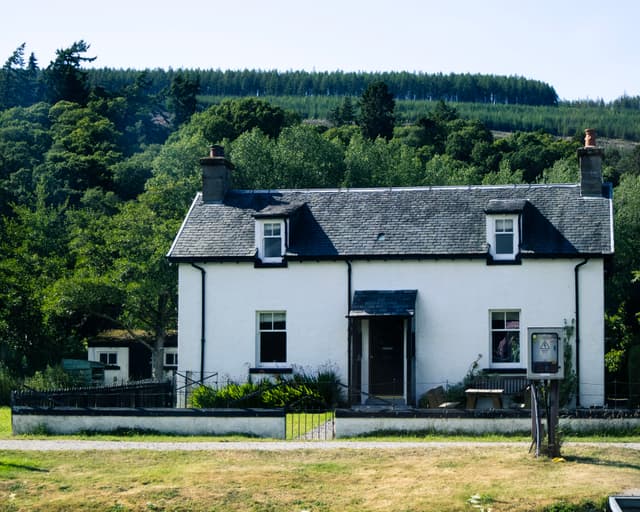 A white two-story cottage with a dark roof, fronted by a trimmed lawn, set against a backdrop of lush trees on a hill