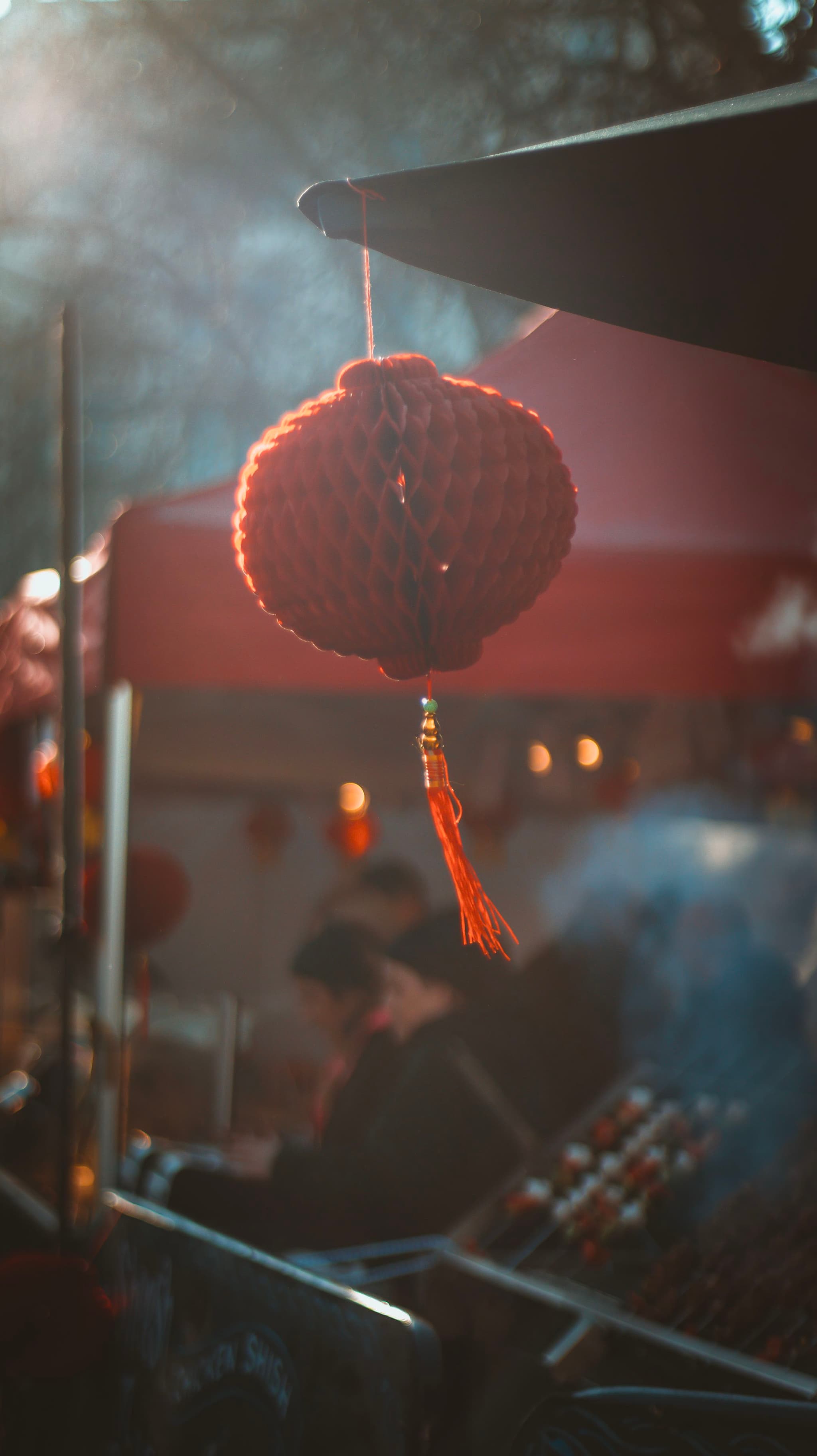 A red lantern hangs in focus against a blurred background of a street food stall with people and ambient smoke, creating a moody, atmospheric setting