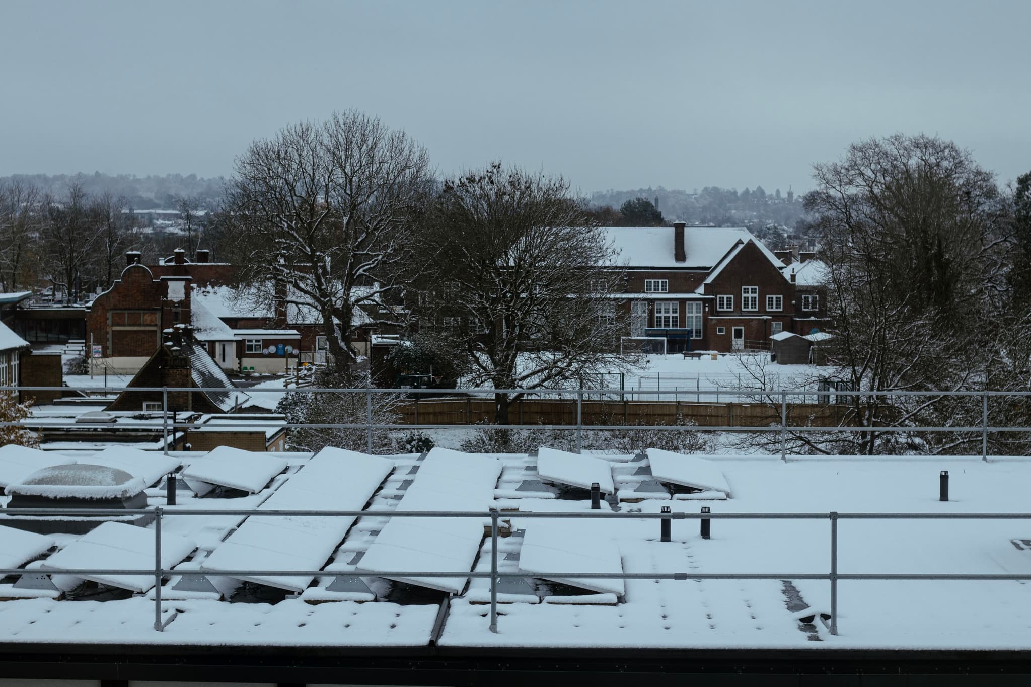 A snow-covered urban landscape including rooftops, trees, and residential buildings, under an overcast sky