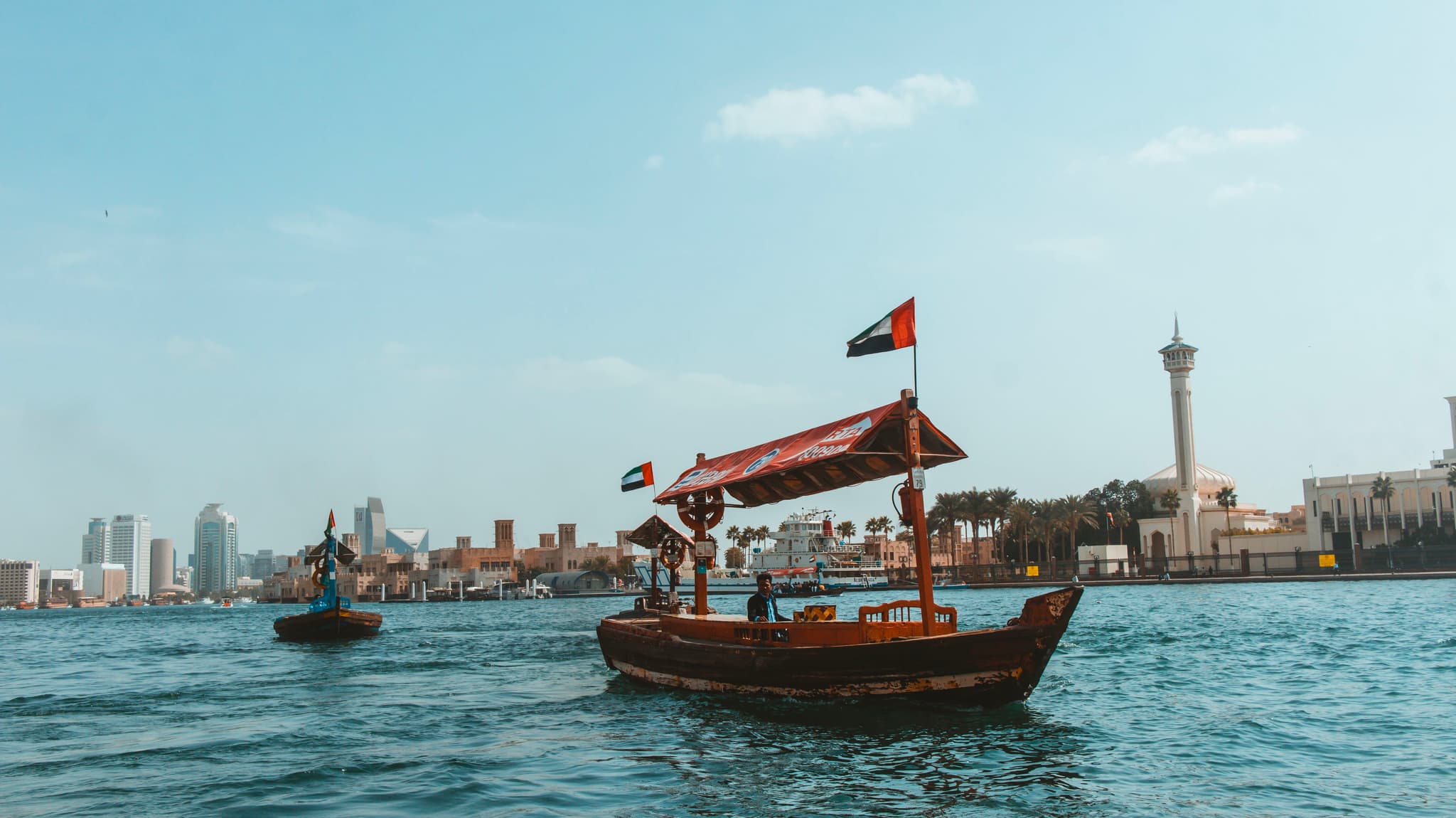 A traditional boat sails on the water with a city skyline and a lighthouse in the background, under a clear blue sky