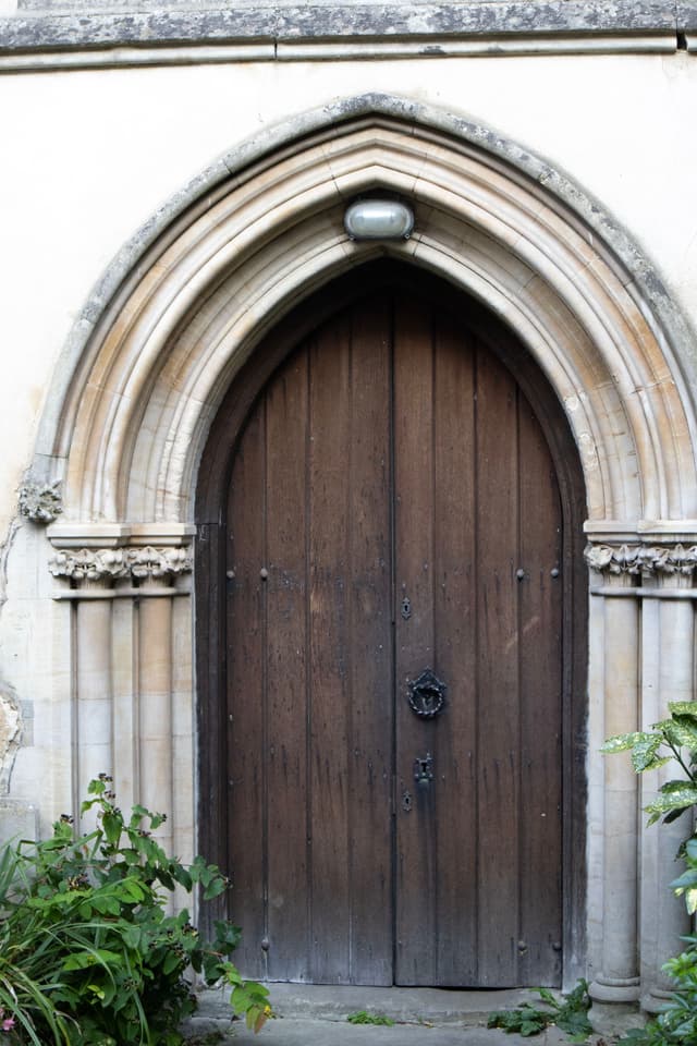 A wooden arched door set within a stone Gothic archway, flanked by some greenery