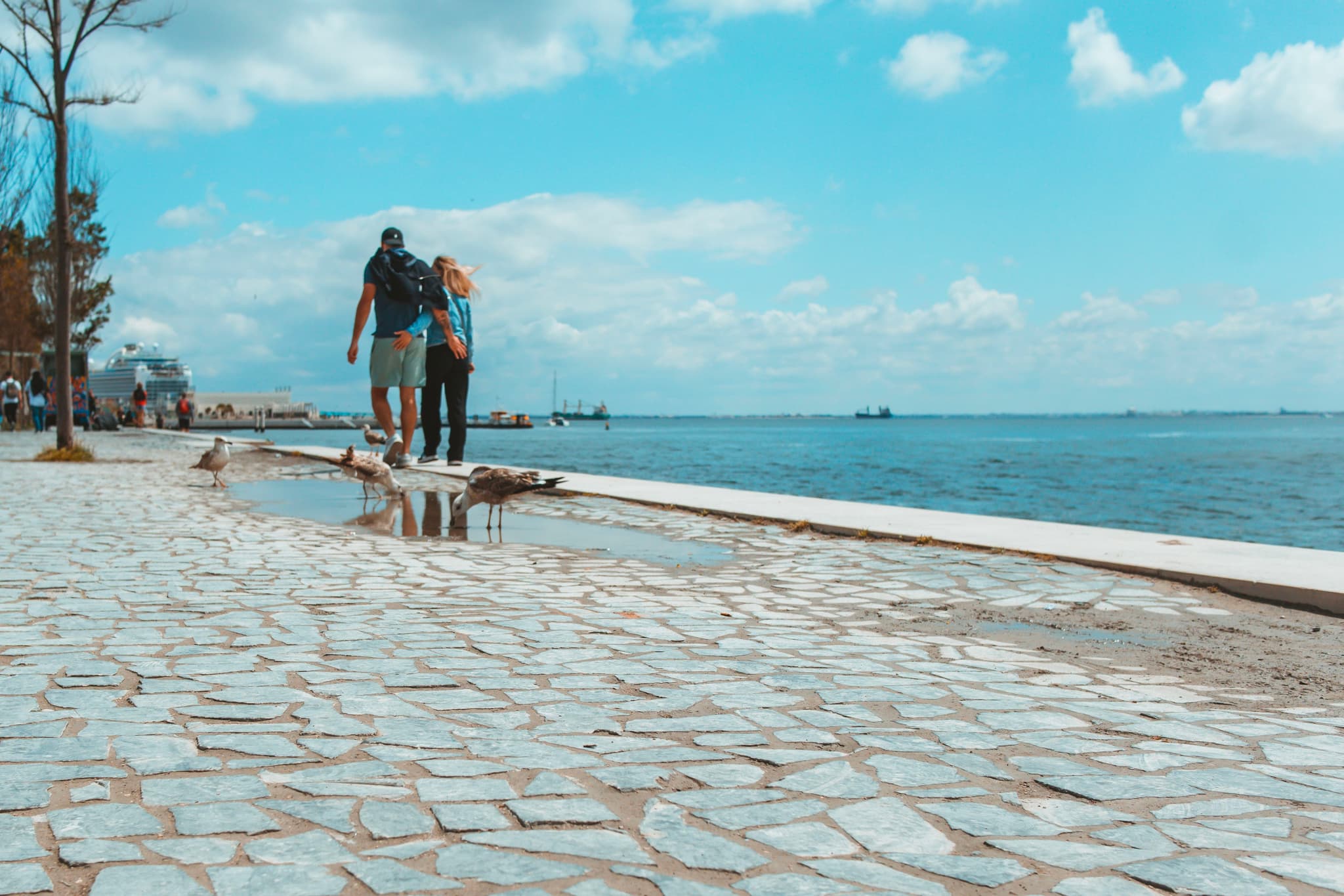 A couple walking along a cobblestone path by the sea under a blue sky with scattered clouds