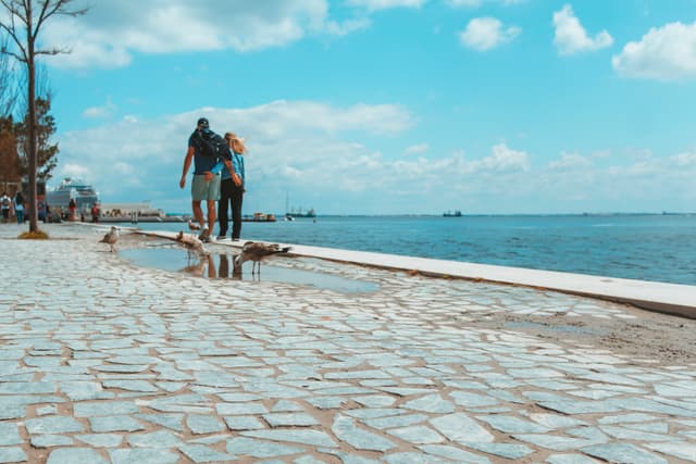 A couple walking along a cobblestone path by the sea under a blue sky with scattered clouds