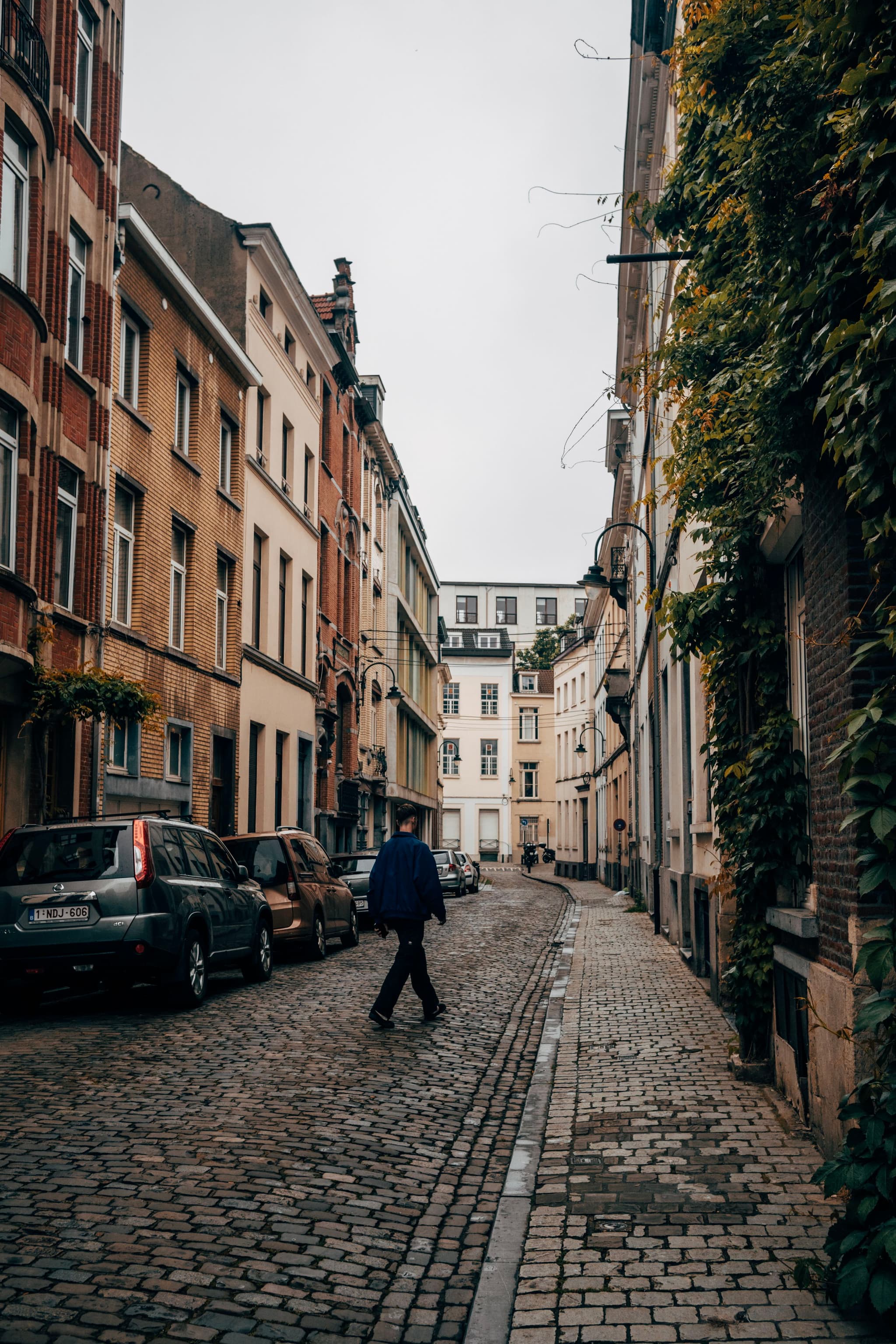 A narrow cobblestone street lined with parked cars and tall buildings, with a person walking in the middle of the street