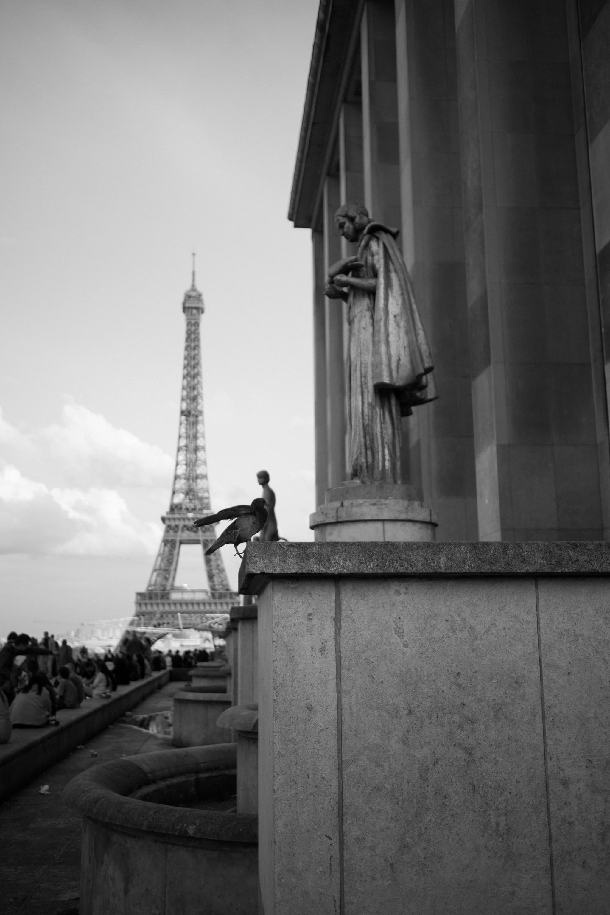 A black and white photo capturing the Eiffel Tower in the background with a person sitting on the ledge of a building, and a statue is visible on the right. It appears to be taken from a high vantage point overlooking the city