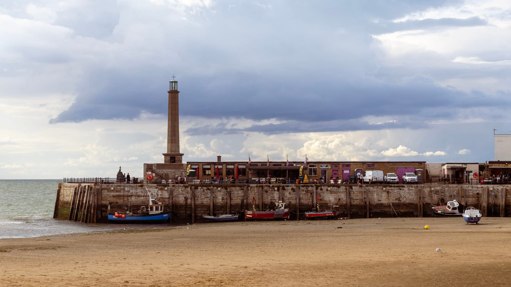 A lighthouse stands on a pier against a cloudy sky, overlooking a beach with a few individuals and boats near the water's edge