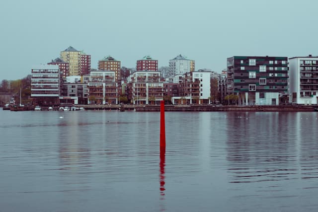 Modern buildings line the waterfront, with a calm body of water in the foreground and a red pole standing vertically in the water - STOCKHOLM SWEDEN