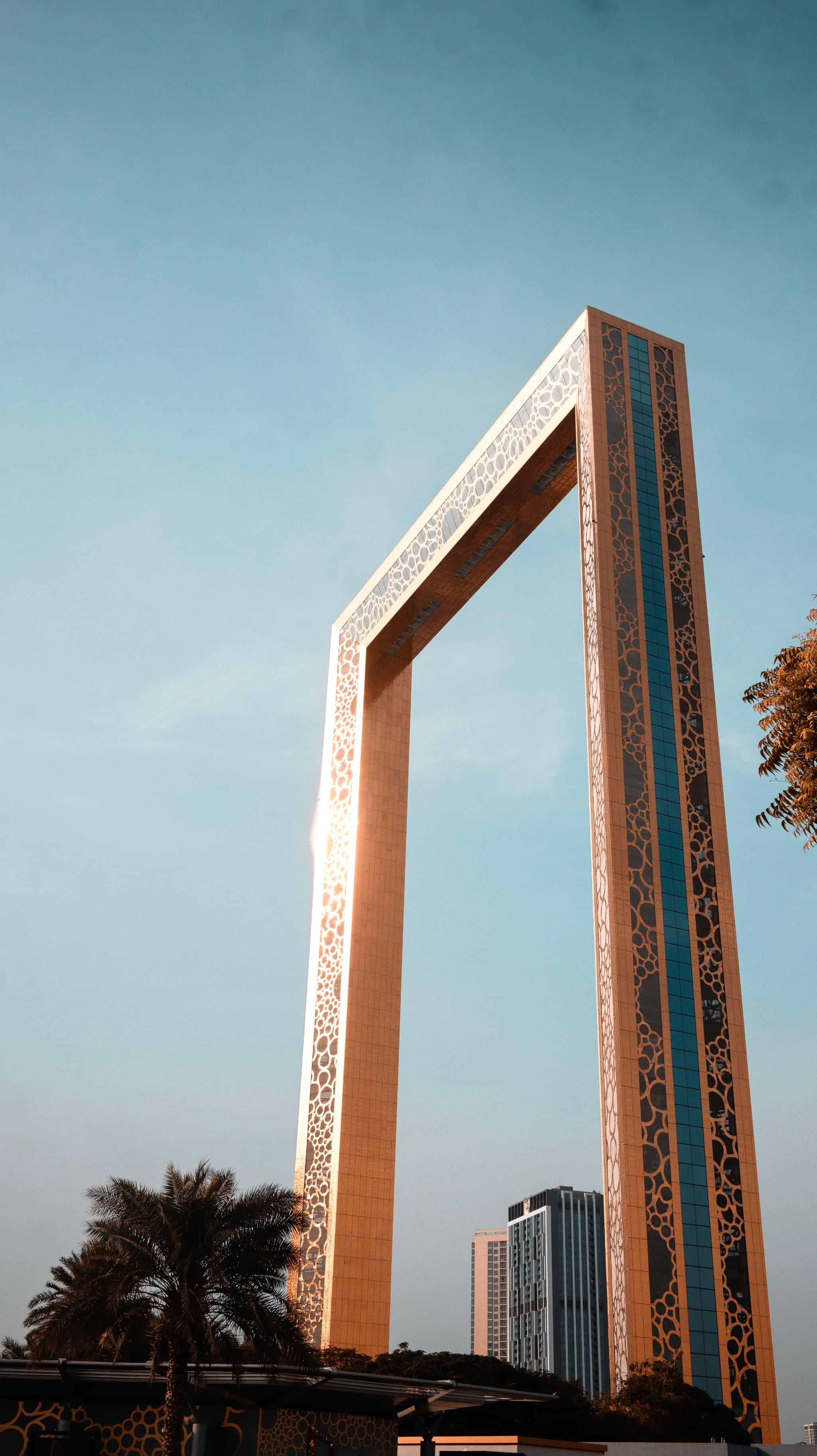 A tall, rectangular arch-like structure with intricate patterns stands against a clear sky, flanked by trees and a distant building