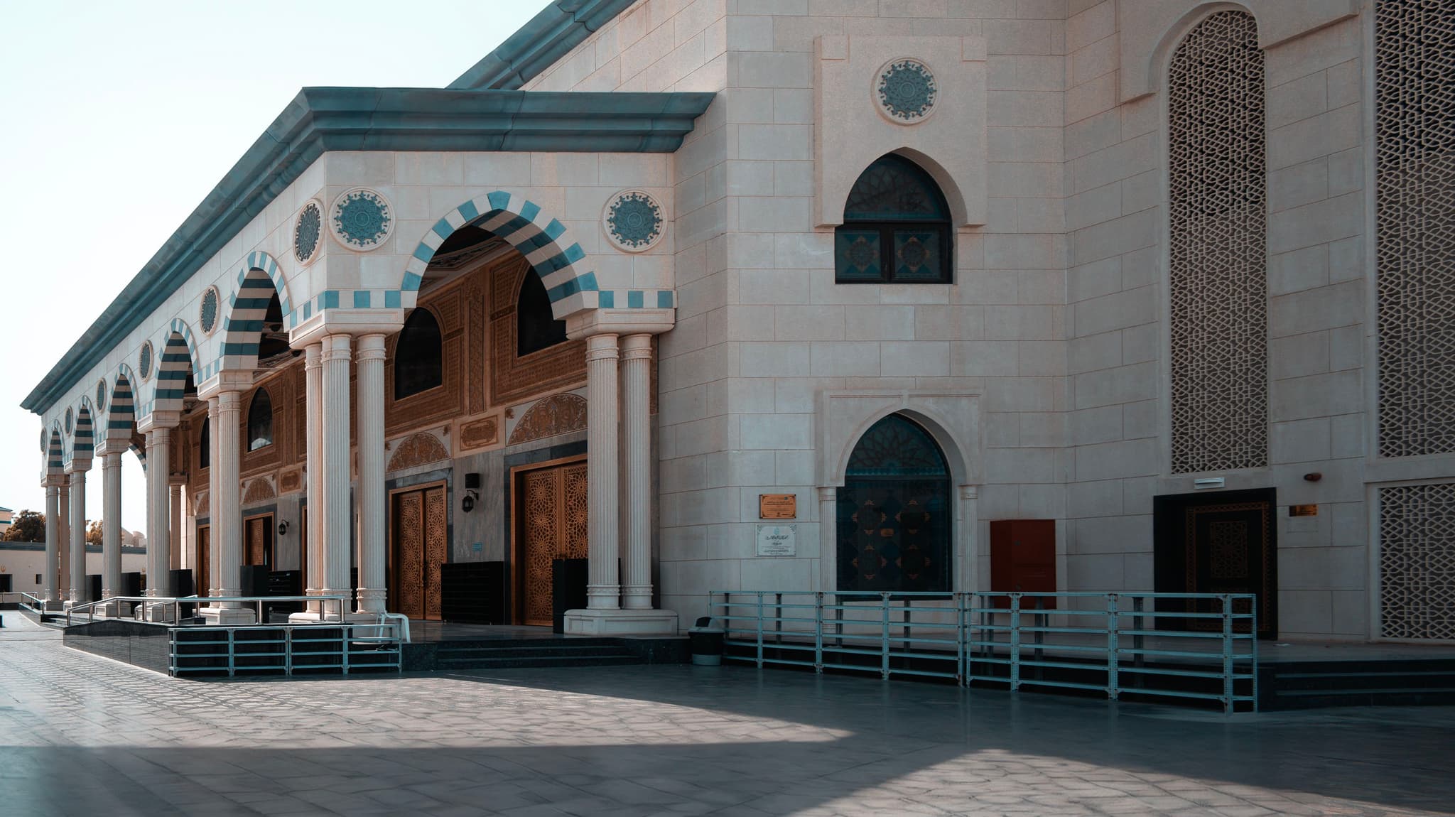 A mosque with a distinctive Islamic architectural style, featuring archways, geometric patterns, and a series of domes and minarets, stands under a clear sky