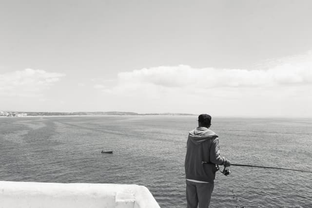 A person fishing while standing on a ledge, overlooking a vast body of water with a small boat in the distance under a cloudy sky
