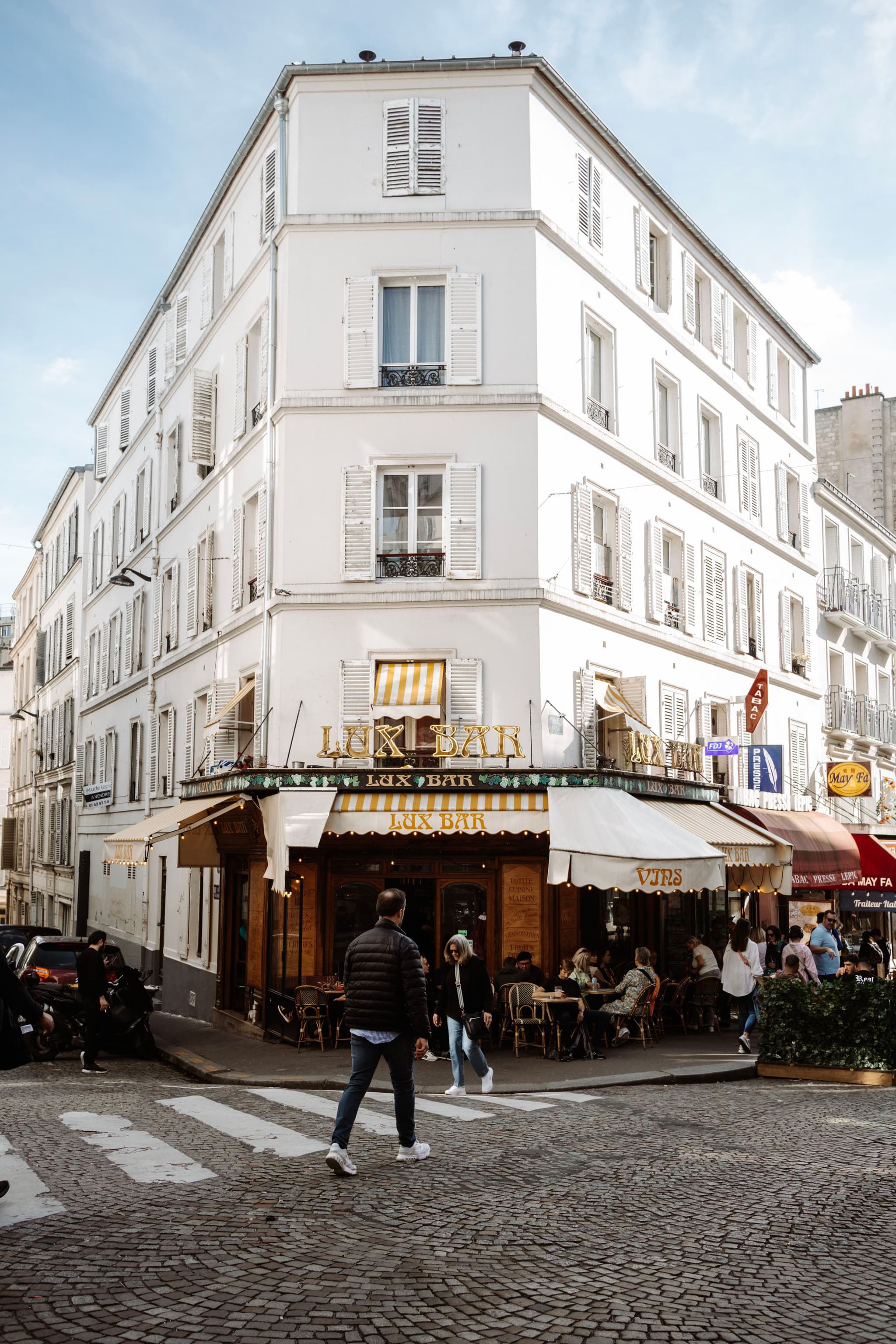 A European-style corner building with a ground-floor café and outdoor seating, under a clear sky, with a man crossing the street in the foreground