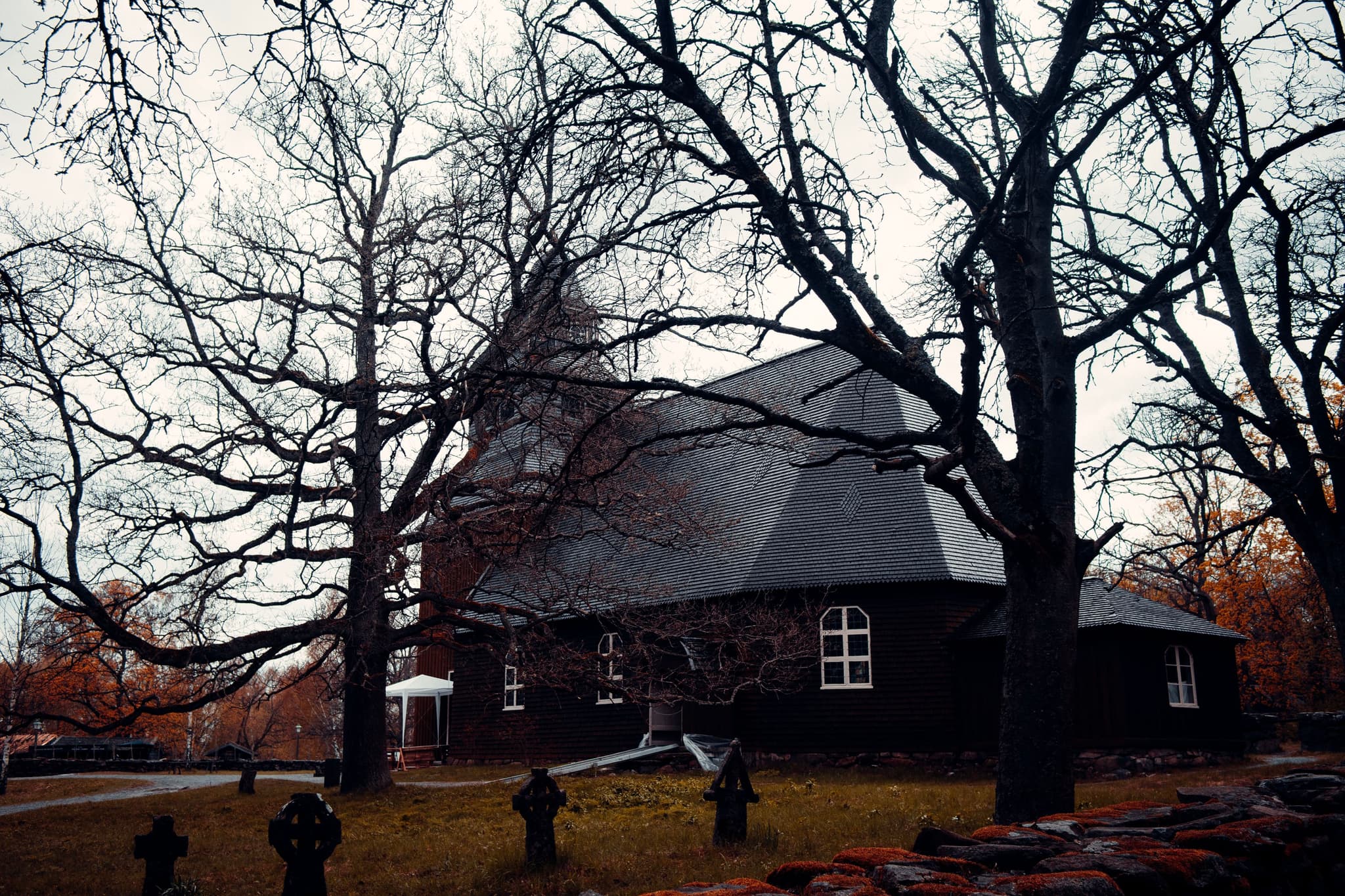 A gloomy, overcast sky looms over a dark, traditional house surrounded by bare trees and fallen leaves, creating an eerie, autumnal scene