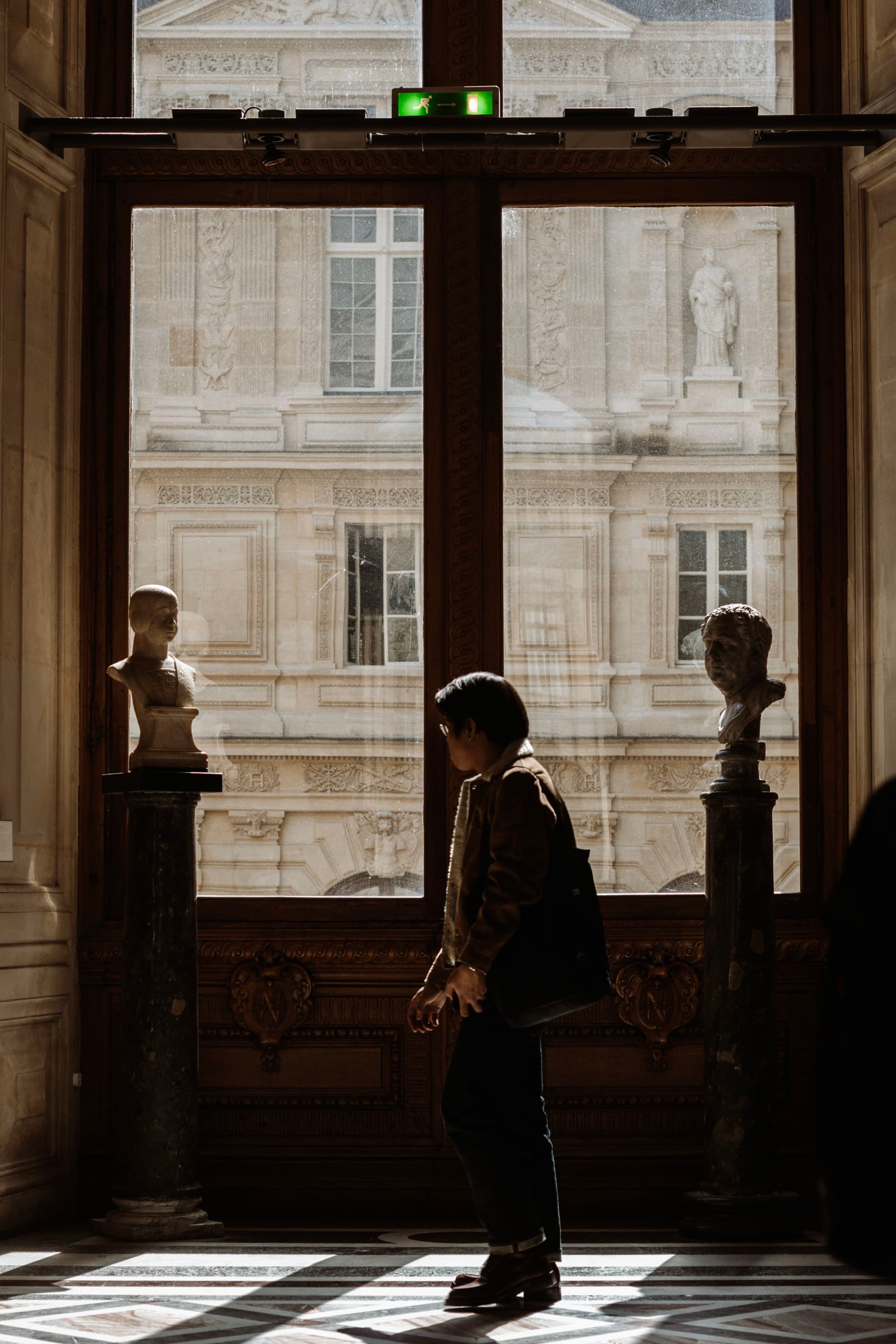 A person stands in a sunlit corridor near an open window, with classical architecture and sculptures visible outside. Shadows create a contrast between the light and dark areas of the scene