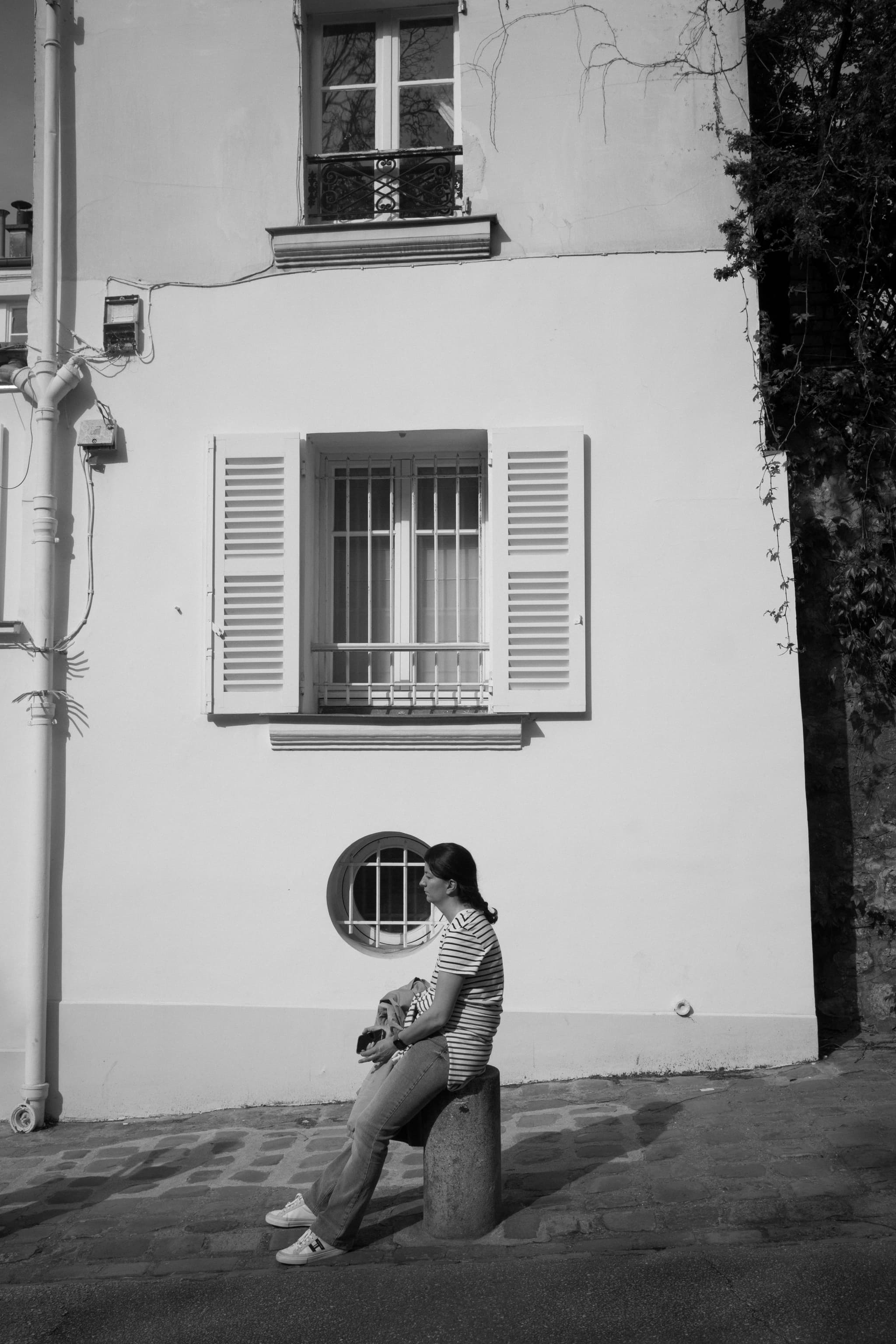 A black and white photograph capturing a person sitting by a circular window on a sunlit street, holding what appears to be a camera, against a backdrop of a building with shutters